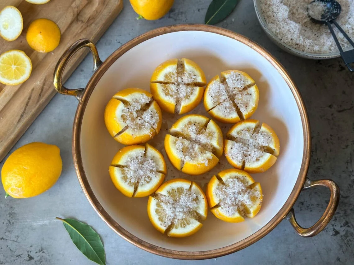 Measured kosher salt, whole lemons, bay leaf, and peppercorns arranged on a light kitchen counter
