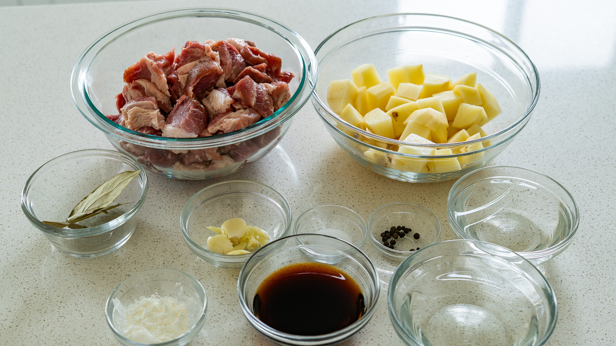 Measured pork adobo ingredients arranged on a tidy kitchen counter: cubed pork shoulder, soy sauce, white vinegar, garlic, bay leaves, whole peppercorns, onion, sugar, and water