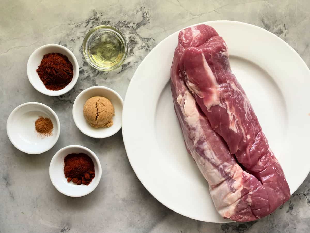 Measured spices and brown sugar in small bowls next to a raw pork loin on a rimmed baking sheet