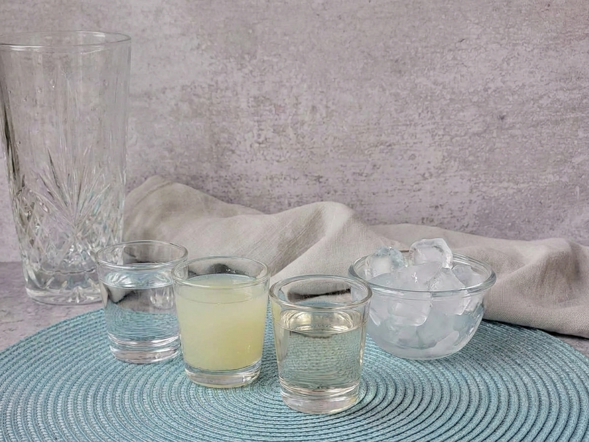 Measured vodka, freshly squeezed lime juice, and simple syrup in small glass cups beside a bowl of ice on a kitchen counter