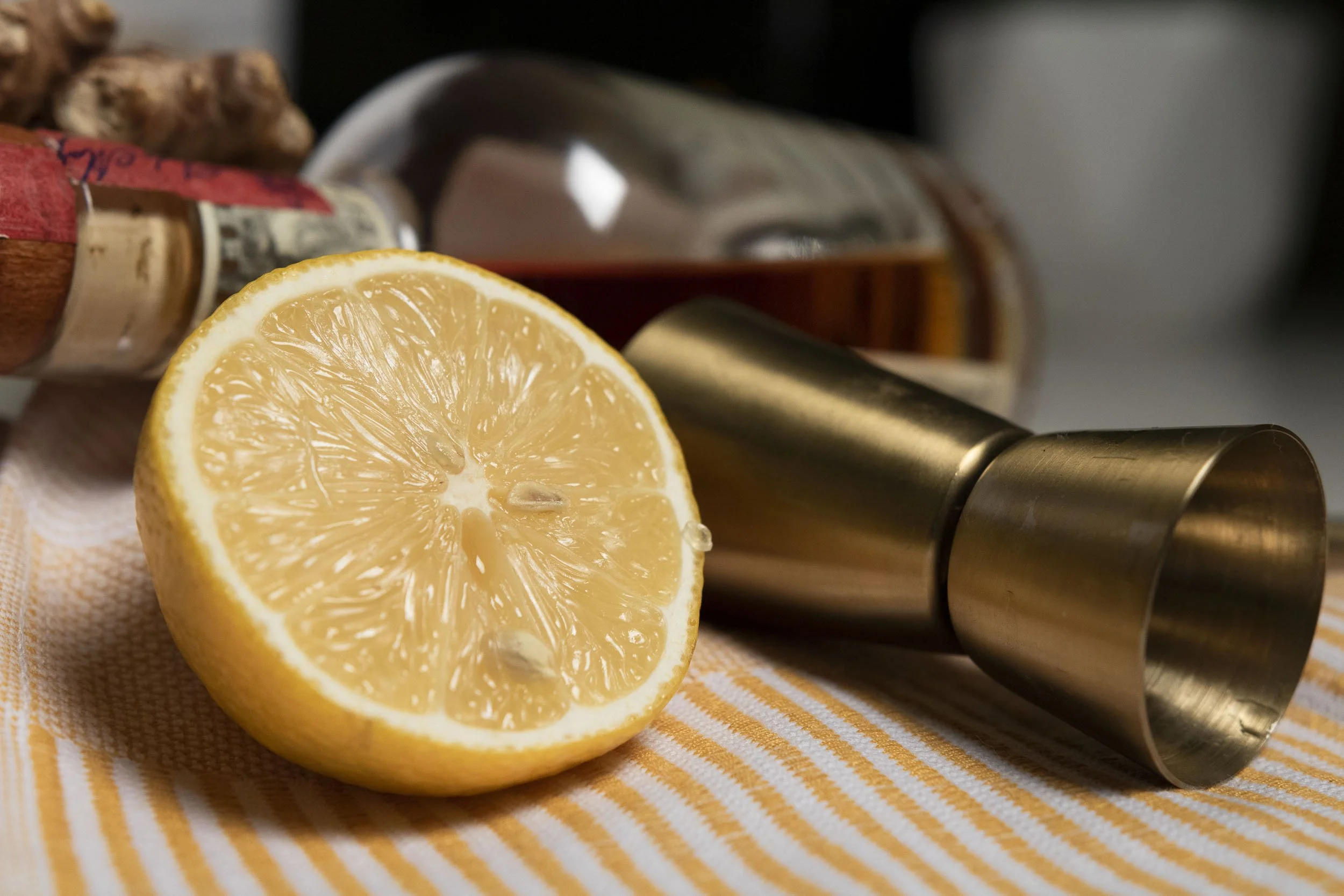 Measured whiskey in a small jigger beside a halved lemon and a spoonful of honey on a wooden cutting board in a home kitchen