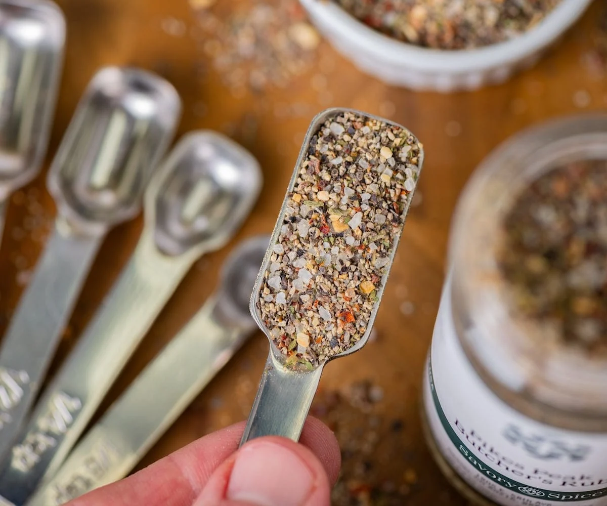 Measuring spoons filled with dried oregano, garlic powder, onion powder, and black pepper arranged on a wooden cutting board on a kitchen counter