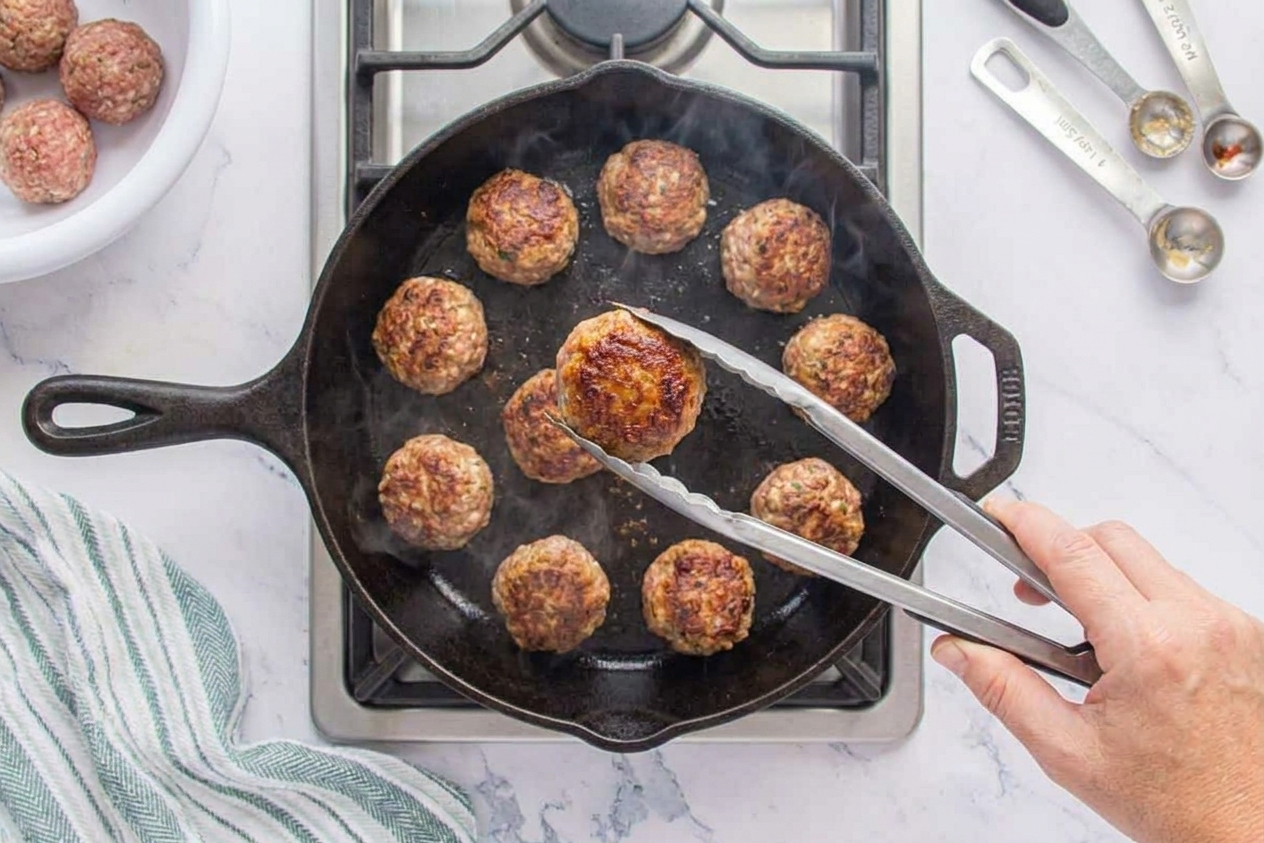Meatballs browning in a cast iron skillet with visible golden crust, held with tongs over a stovetop