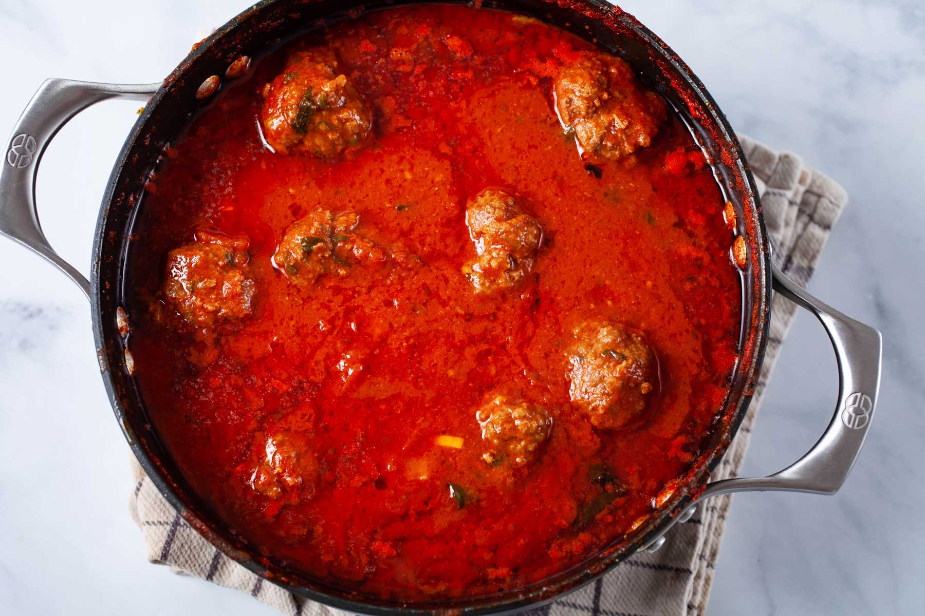 Meatballs gently simmering in a Dutch oven of tomato sauce with small bubbles breaking on the surface on a stovetop