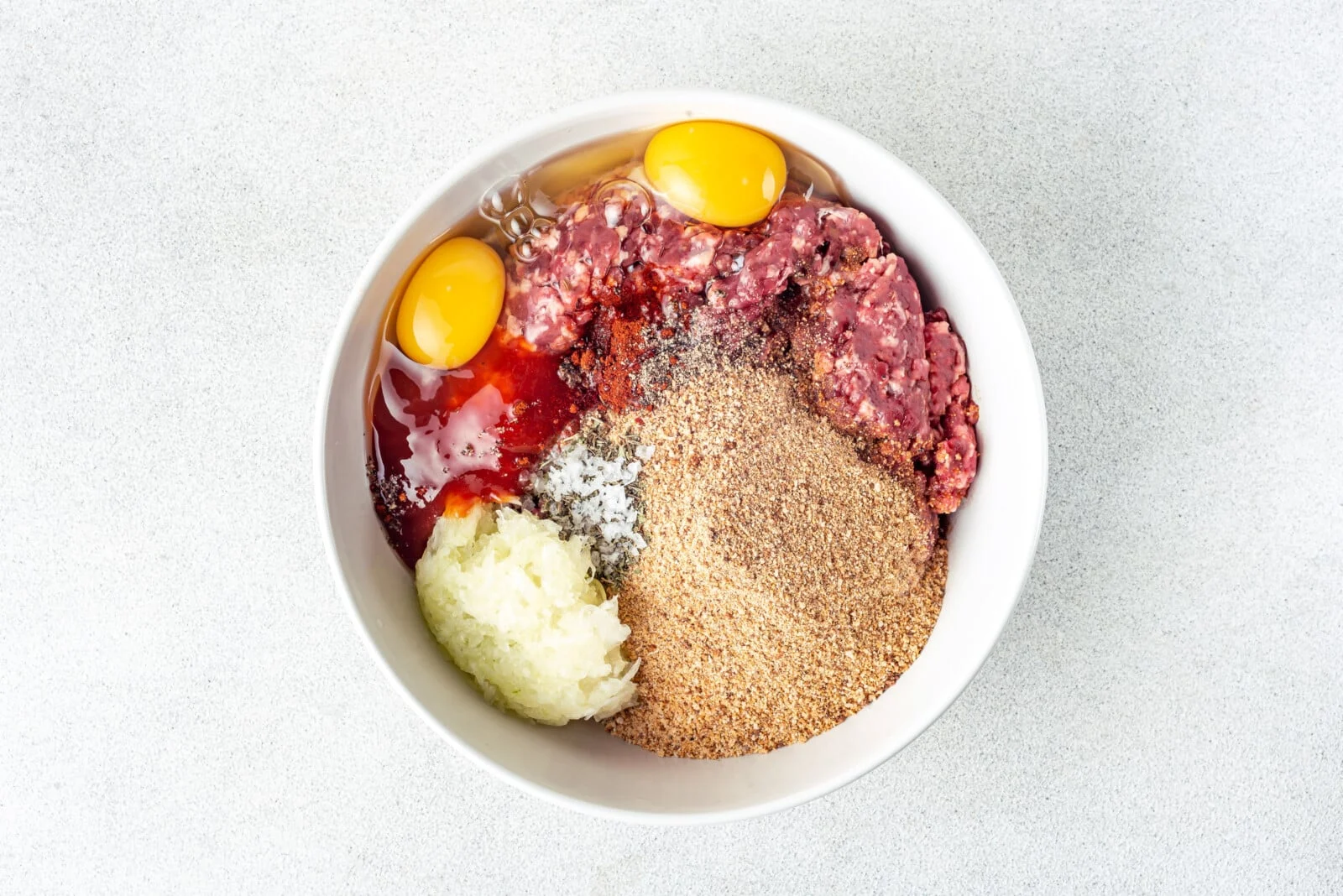 Meatloaf ingredients arranged on a kitchen counter, including ground beef, breadcrumbs, eggs, onion, milk, ketchup, brown sugar, and spices in small bowls.