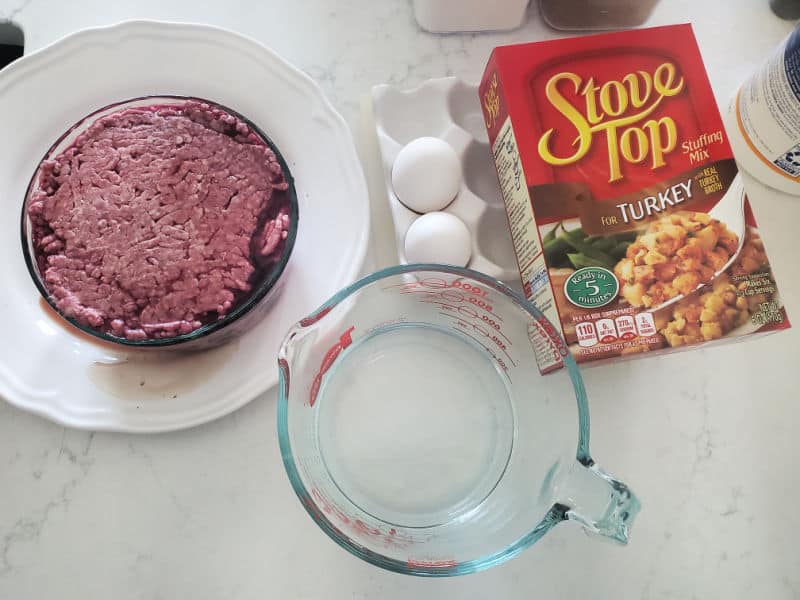 Meatloaf ingredients including ground beef, stuffing mix, eggs, milk, onion, garlic, ketchup, mustard, brown sugar, and shredded cheese arranged together on a kitchen counter