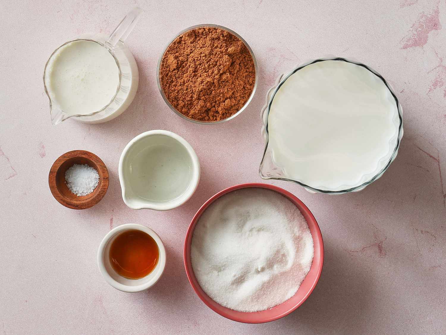 Milk, cocoa powder, sugars, vanilla, and salt arranged on a bright kitchen counter with a small saucepan nearby