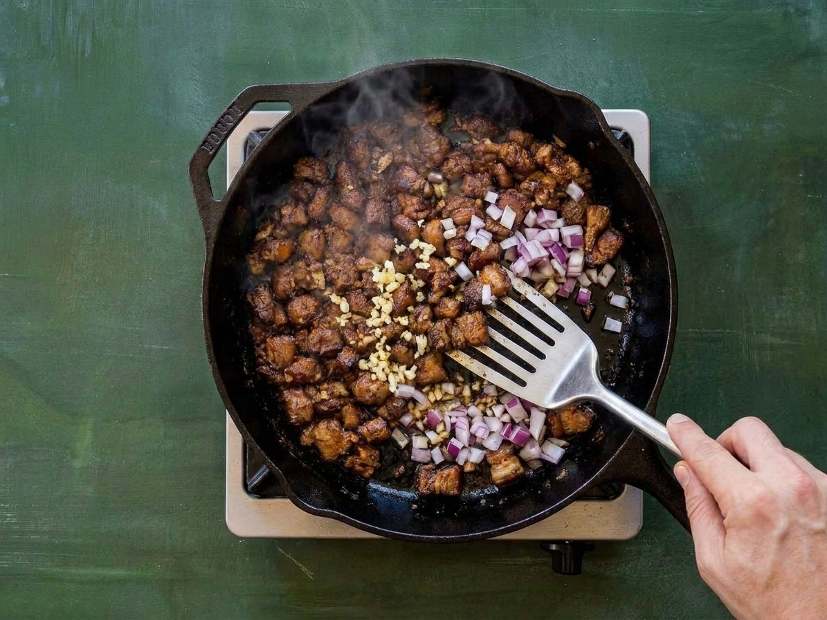 Minced garlic and diced red onion being sautéed with browned pork in a skillet on a stovetop