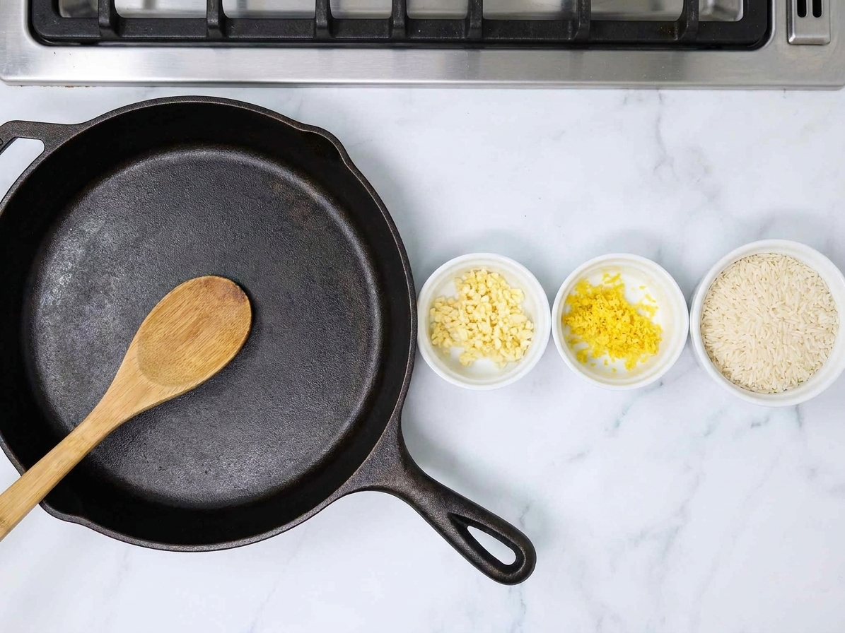 Minced garlic, lemon zest, and uncooked long grain rice in small bowls next to a large skillet on a kitchen counter