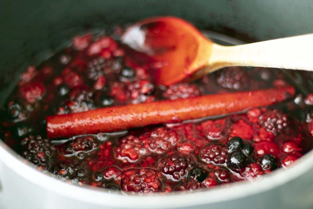Mixed berries simmering into a glossy compote in a small saucepan on a stovetop with a wooden spoon nearby