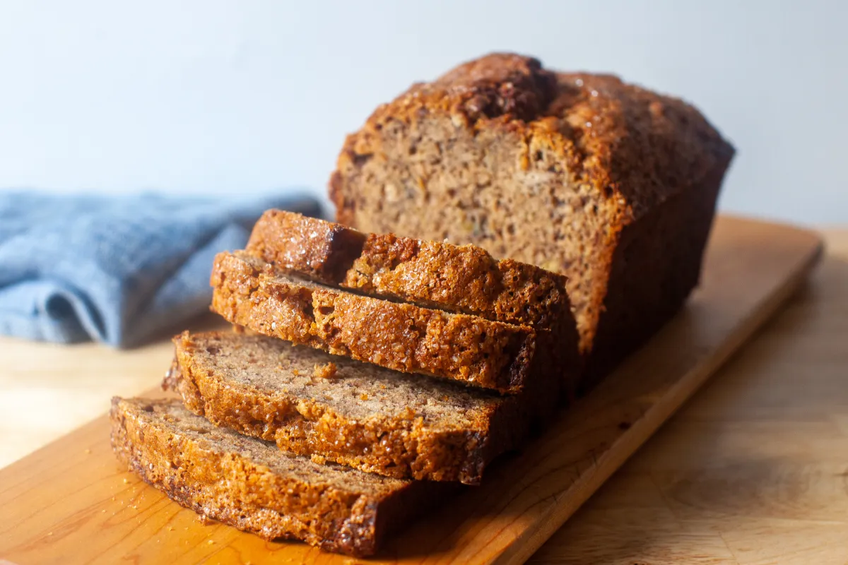 Moist banana bread loaf on a wooden cutting board with one thick slice cut to show a tender crumb with banana specks, lit by natural window light