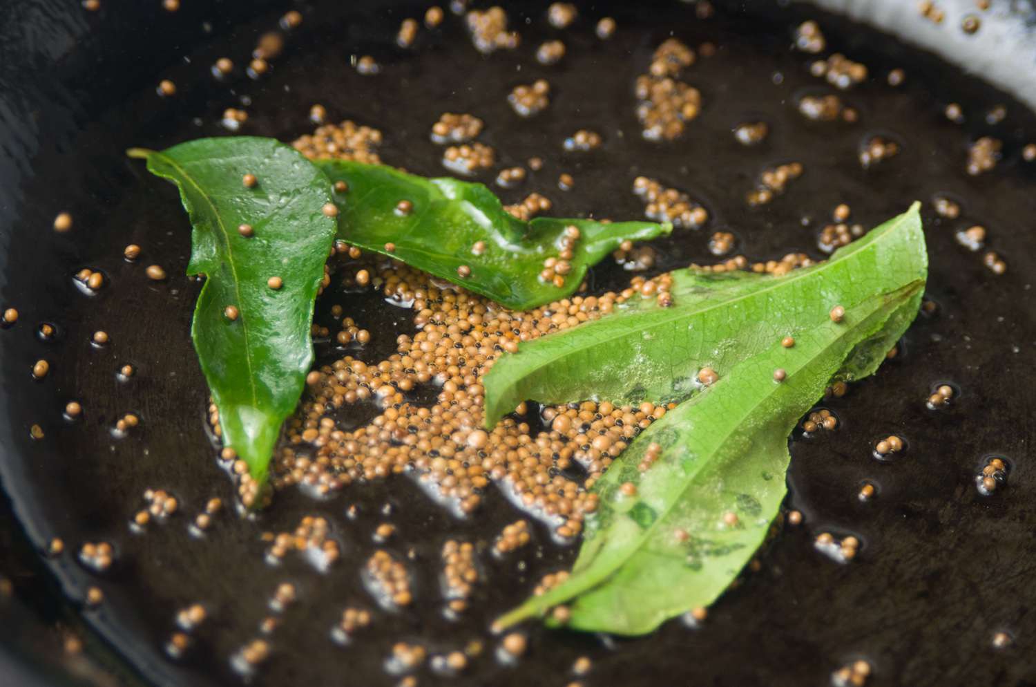 Mustard seeds popping in hot oil with curry leaves in a small skillet on a stovetop