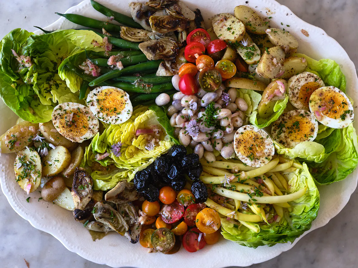 Niçoise salad ingredients arranged on a kitchen counter including eggs, baby potatoes, green beans, cherry tomatoes, canned tuna, olives, capers, lemon, Dijon mustard, and olive oil