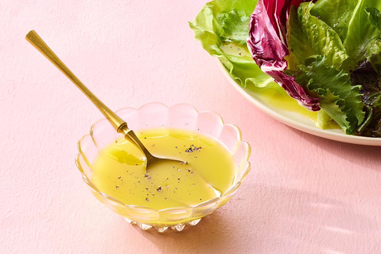 Olive oil being slowly poured into a small bowl with apple cider vinegar and Dijon while a whisk rests on the rim, on a bright kitchen counter