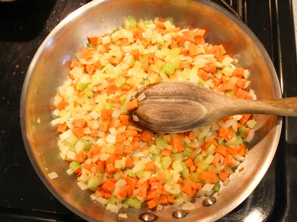 Onion, carrot, and celery sautéing in a large pot on a stovetop with a wooden spoon stirring