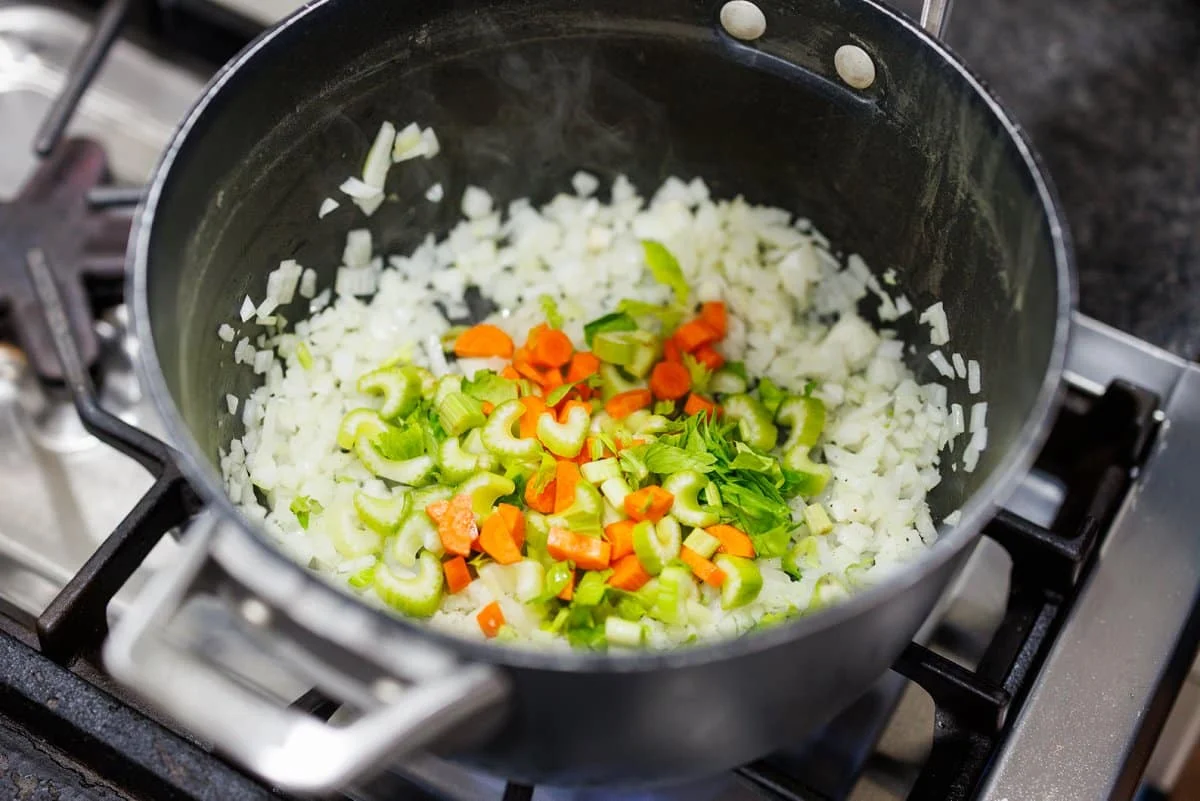Onion, carrot, and celery sautéing in a large soup pot on a stovetop, lightly glossy from butter