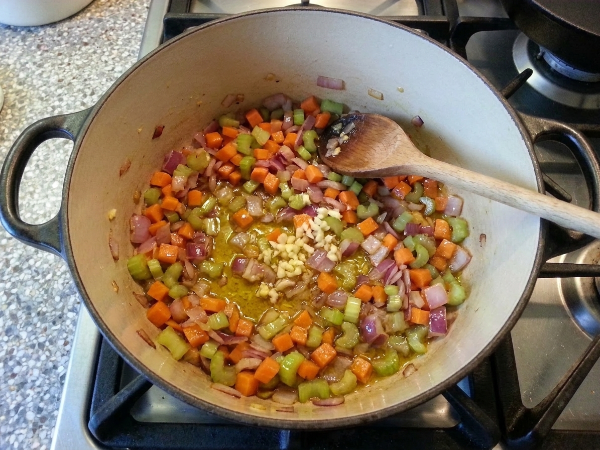 Onion, carrot, celery, and garlic sautéing in a Dutch oven on a stovetop, vegetables softened and glossy