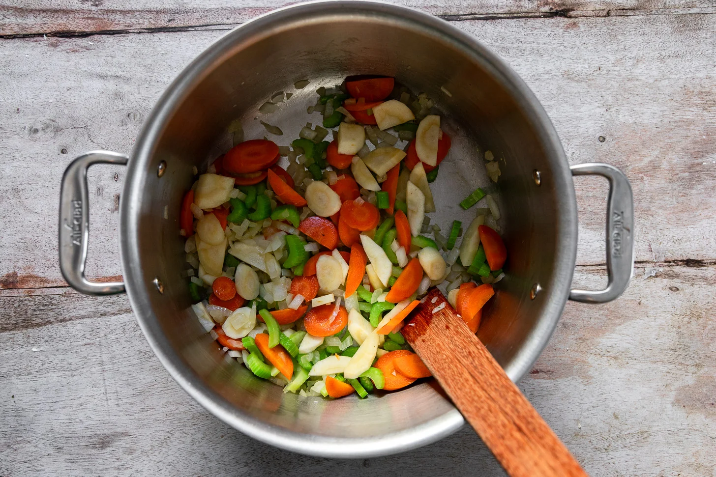 Onion, carrots, celery, and diced apple sautéing in a large Dutch oven on a stovetop with a wooden spoon stirring