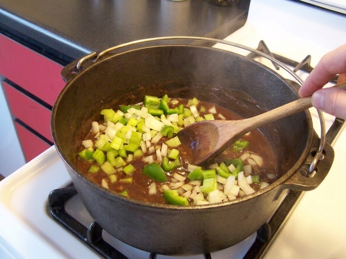 Onion, celery, and green bell pepper cooking in a dark roux inside a Dutch oven, with a spoon stirring