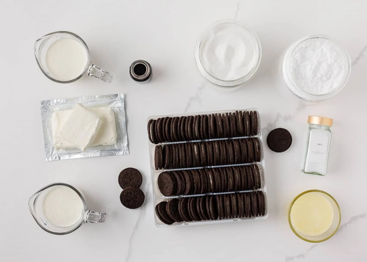 Oreos, blocks of cream cheese, butter, powdered sugar, and a carton of heavy cream arranged on a kitchen counter