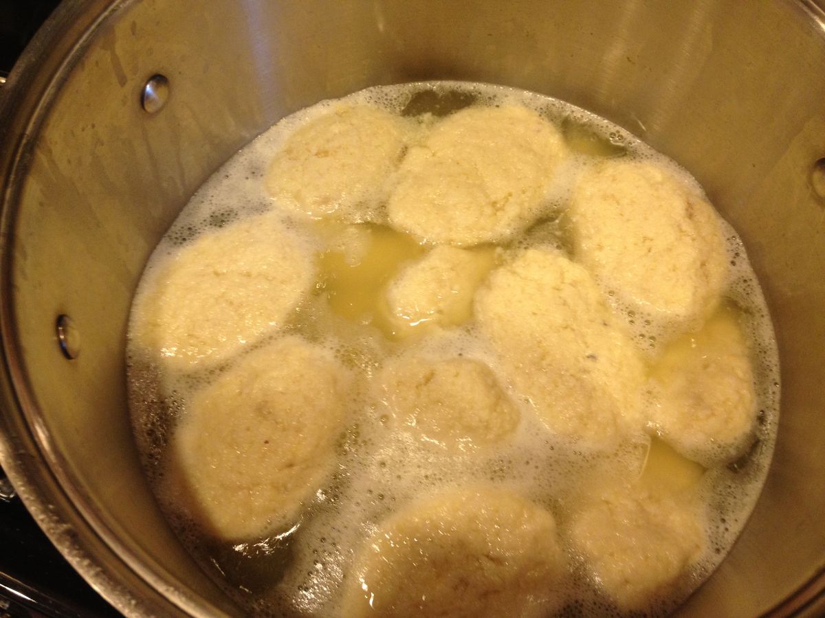 Oval gefilte fish patties gently simmering in a wide pot of clear broth with carrot slices and onion pieces, shot from above on a stovetop