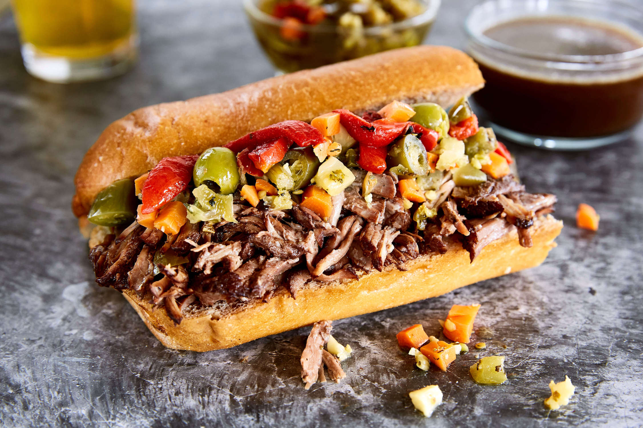 Overhead photo of Italian beef sandwich ingredients on a kitchen counter: chuck roast, beef broth, garlic, Italian seasoning, pepperoncini, onions, bell peppers, rolls, and a jar of giardiniera