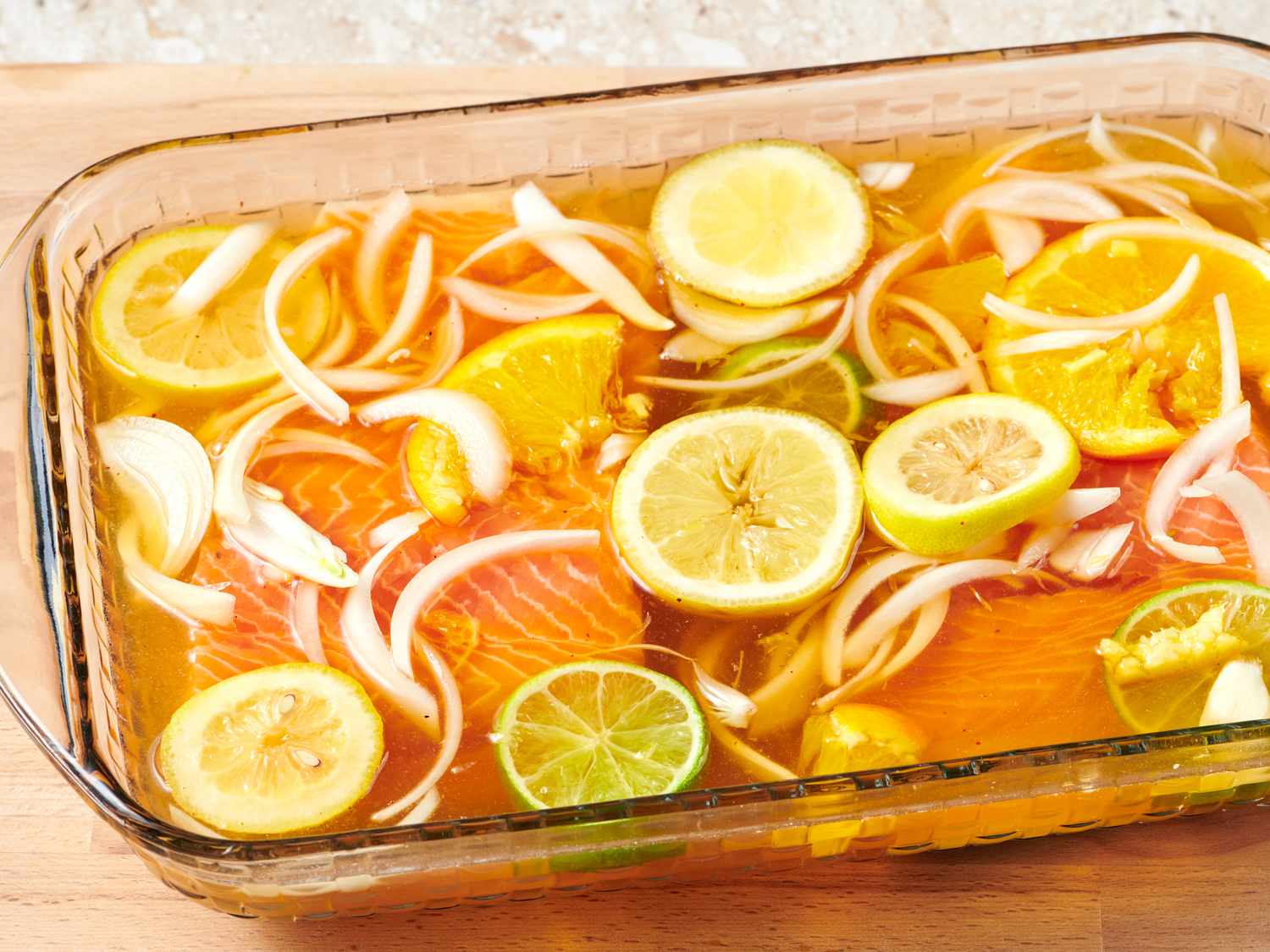 Overhead view of brine ingredients on a light kitchen counter: kosher salt, brown sugar, garlic, black peppercorns, bay leaves, lemon, and a pitcher of water beside raw salmon fillets