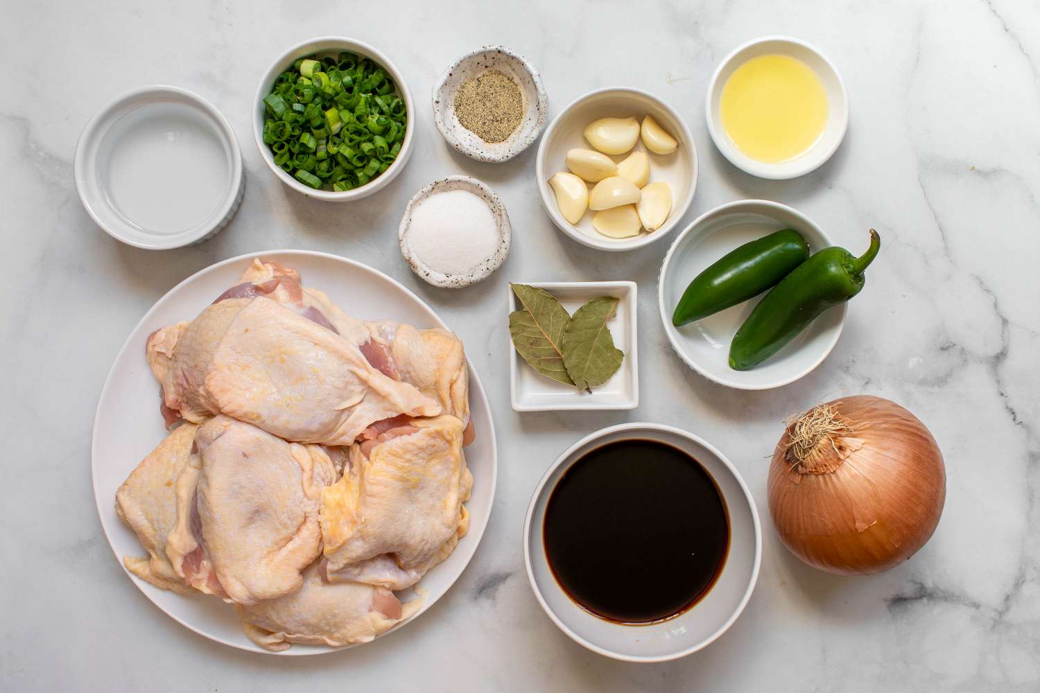 Overhead view of ingredients for chicken adobo on a kitchen counter including chicken thighs, soy sauce, cane vinegar, garlic, bay leaves, black peppercorns, and a small bowl of brown sugar
