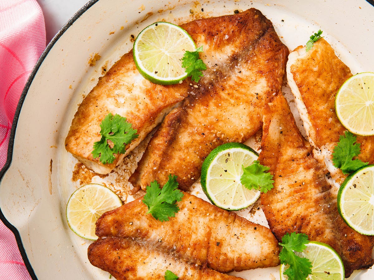 Pan-seared tilapia fillets with golden crust in a skillet with lemon slices and chopped parsley