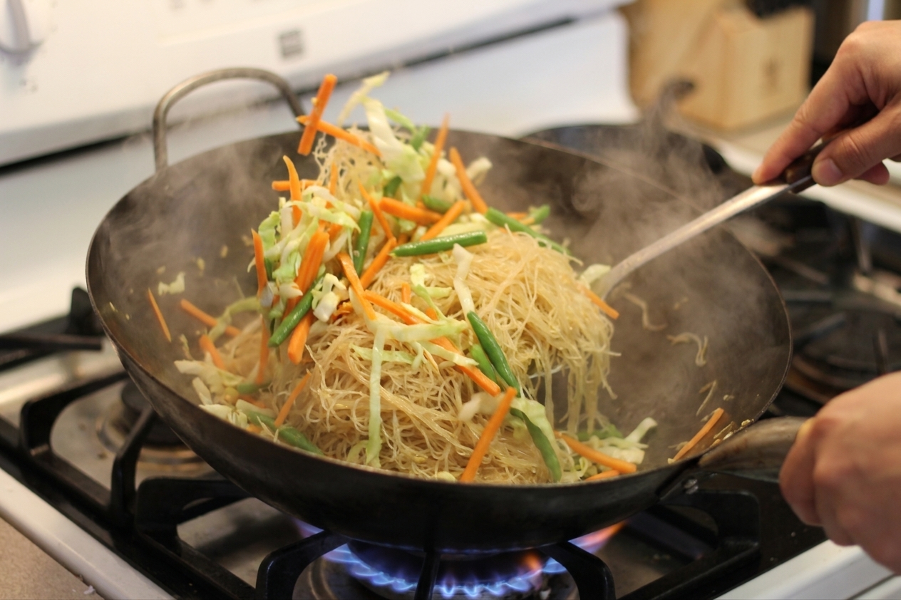 Pancit bihon noodles being tossed with vegetables in a hot wok on a stovetop with steam rising
