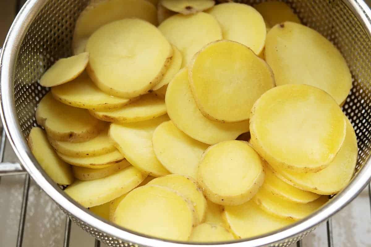 Parboiled potato slices draining in a colander over a sink, steam rising in a home kitchen