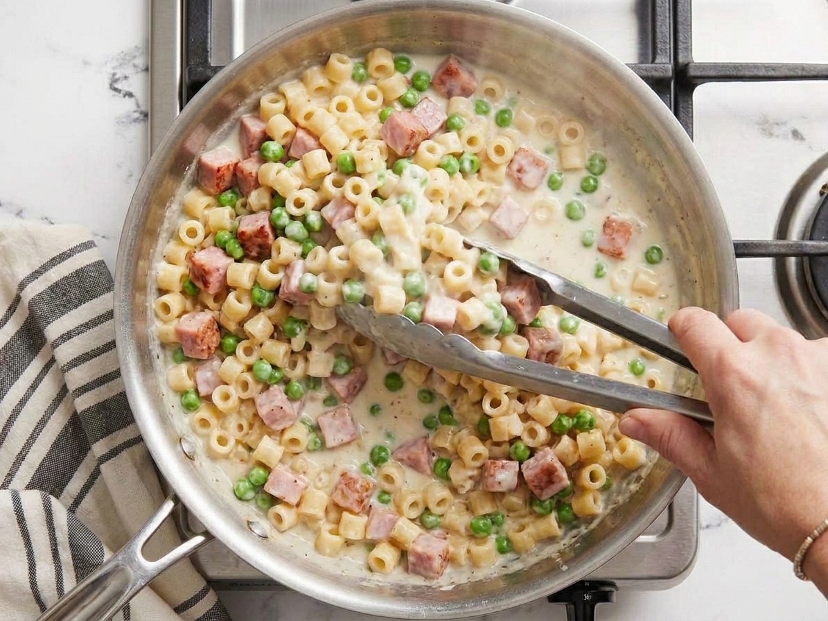 Pasta and peas being tossed in a skillet with creamy ham sauce using tongs on a home stovetop