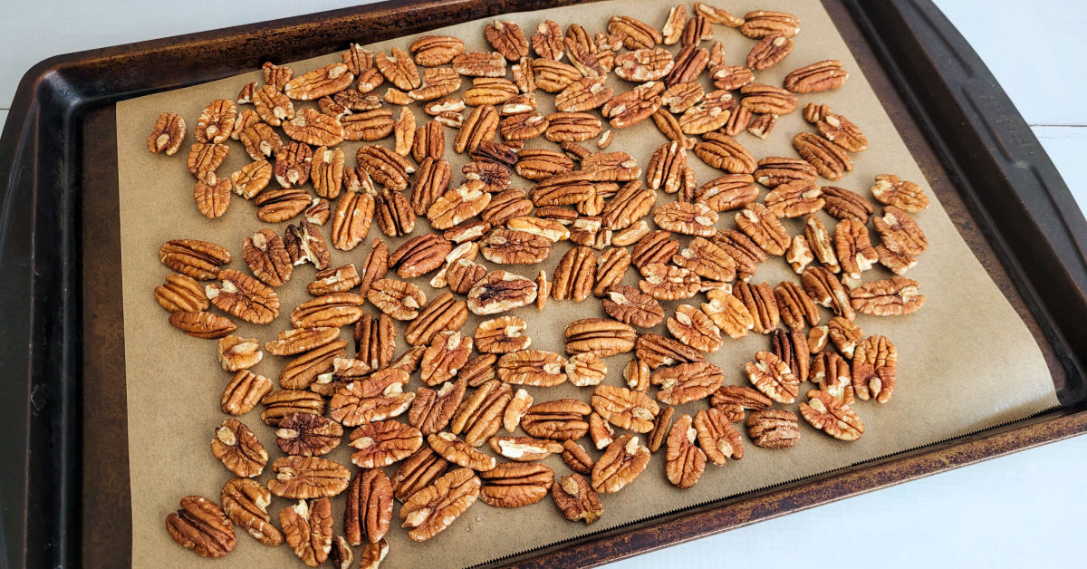 Pecan halves spread on a baking sheet after toasting, golden and fragrant-looking in a warmly lit kitchen