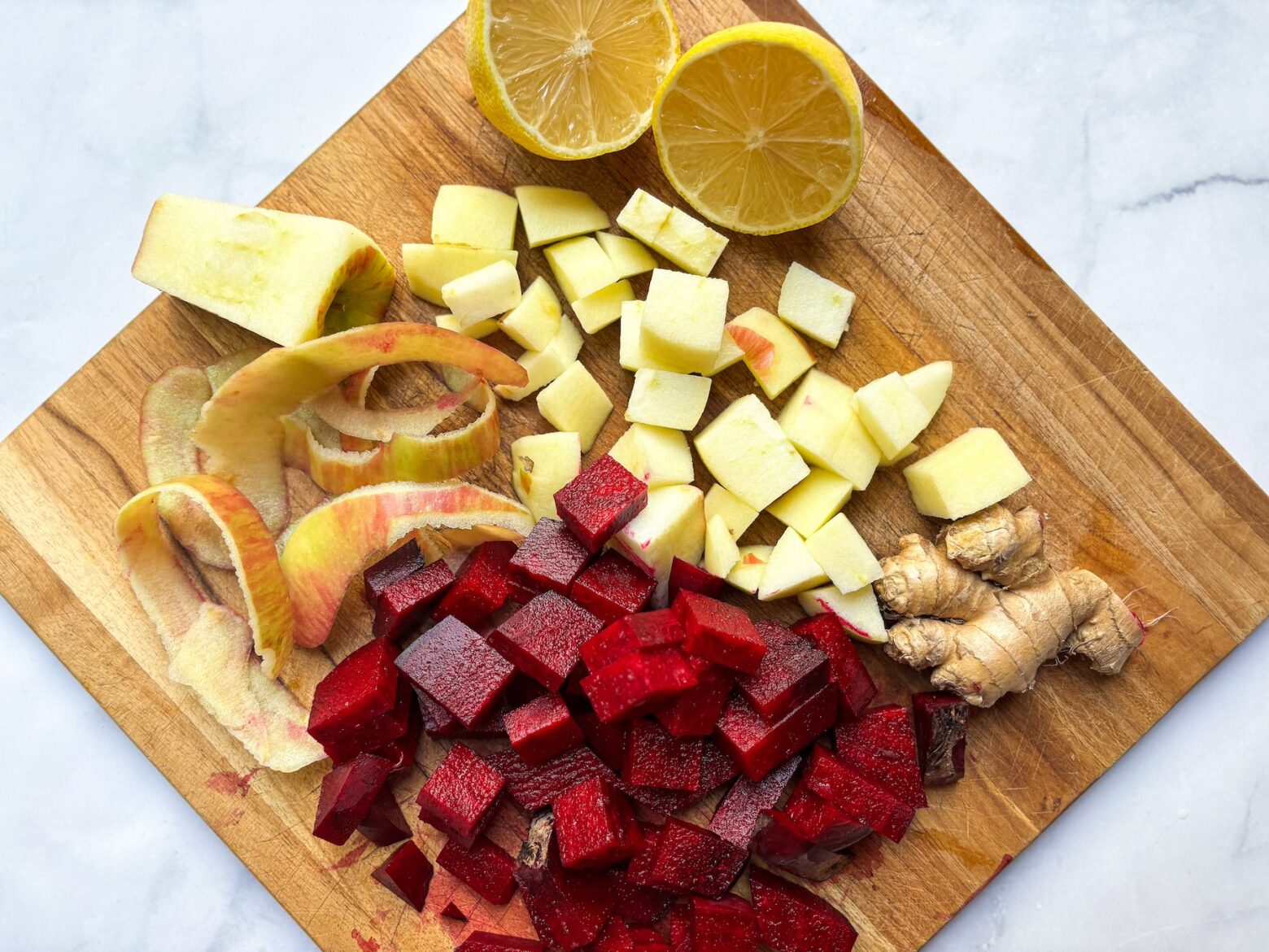 Peeled and chopped beet chunks on a wooden cutting board with a chef's knife, lemon halves, and ginger nearby