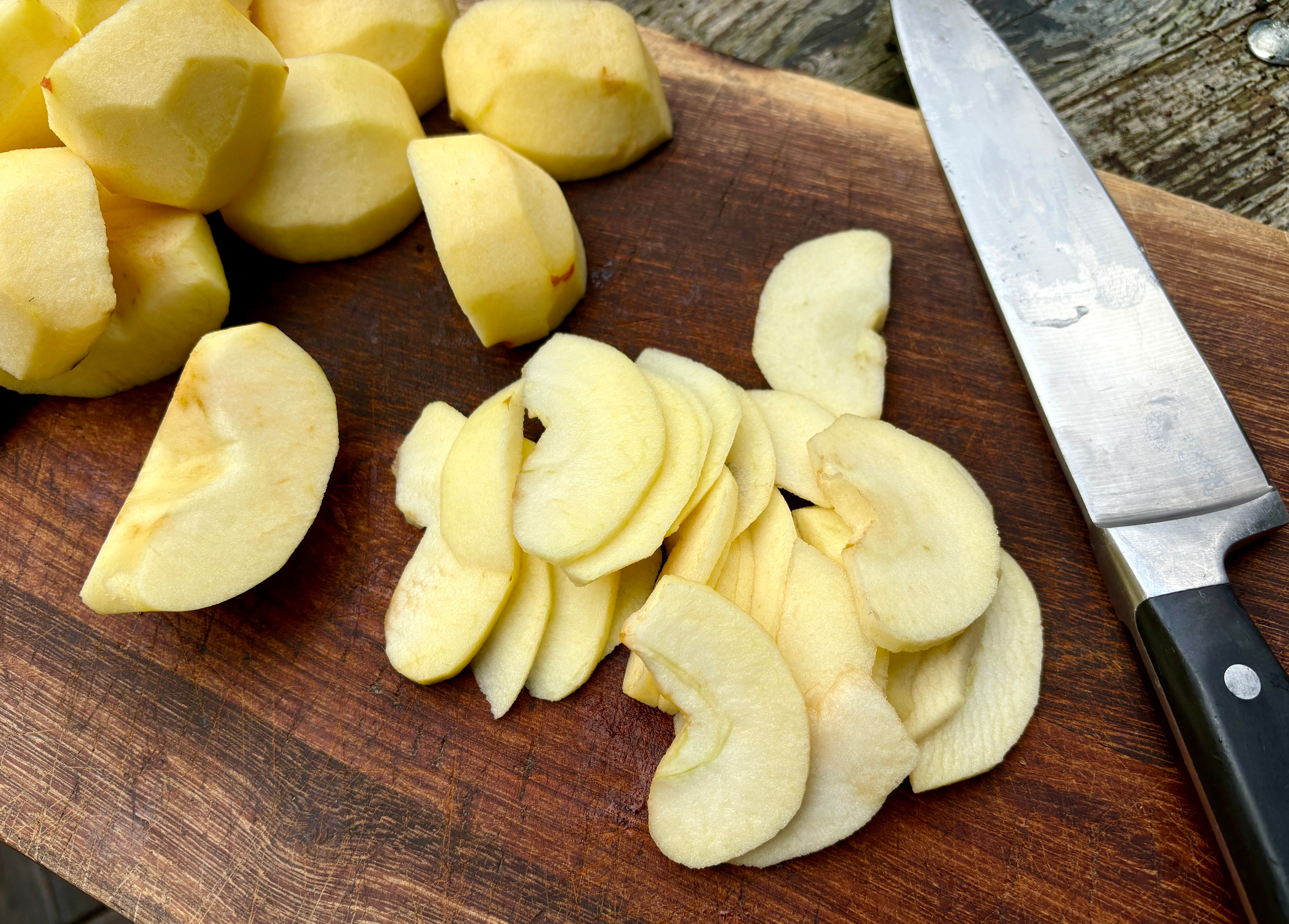 Peeled apples cut into large wedges on a wooden cutting board beside a chef’s knife on a bright kitchen counter
