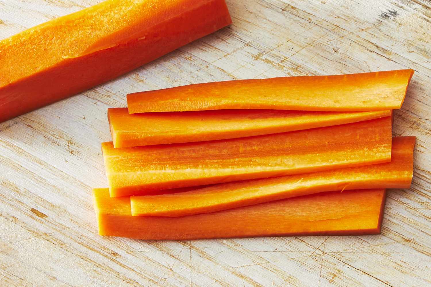 Peeled carrots cut into even sticks on a wooden cutting board with a chef’s knife beside them
