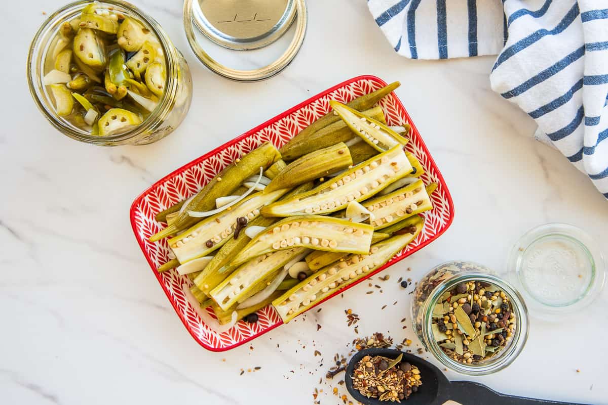 Pickled okra pods served on a small snack board with sliced cheddar, crackers, and a ramekin of mustard on a wooden table.