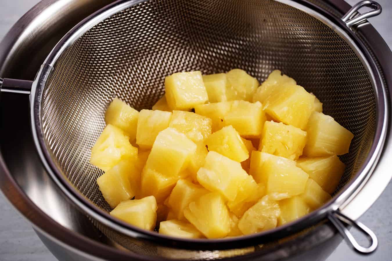 Pineapple chunks draining in a metal colander set over a bowl on a kitchen counter