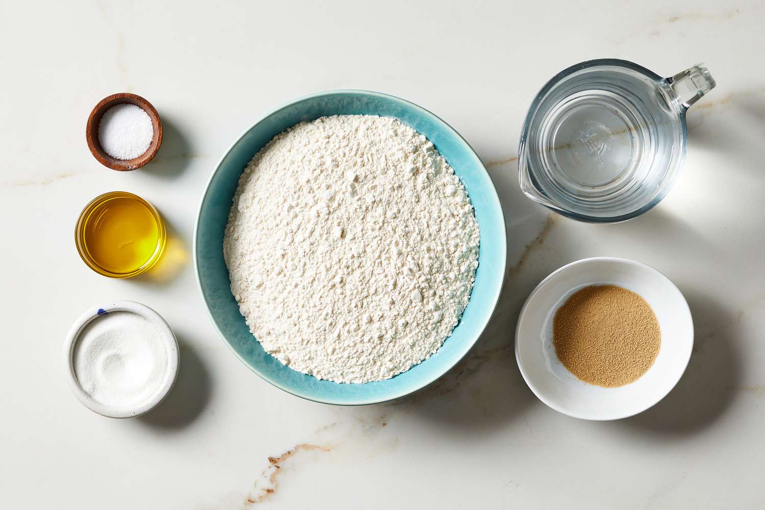 Pizza dough ingredients arranged on a wooden board, including flour in a bowl, a small dish of yeast, a measuring cup of warm water, salt, and olive oil on a cozy kitchen counter