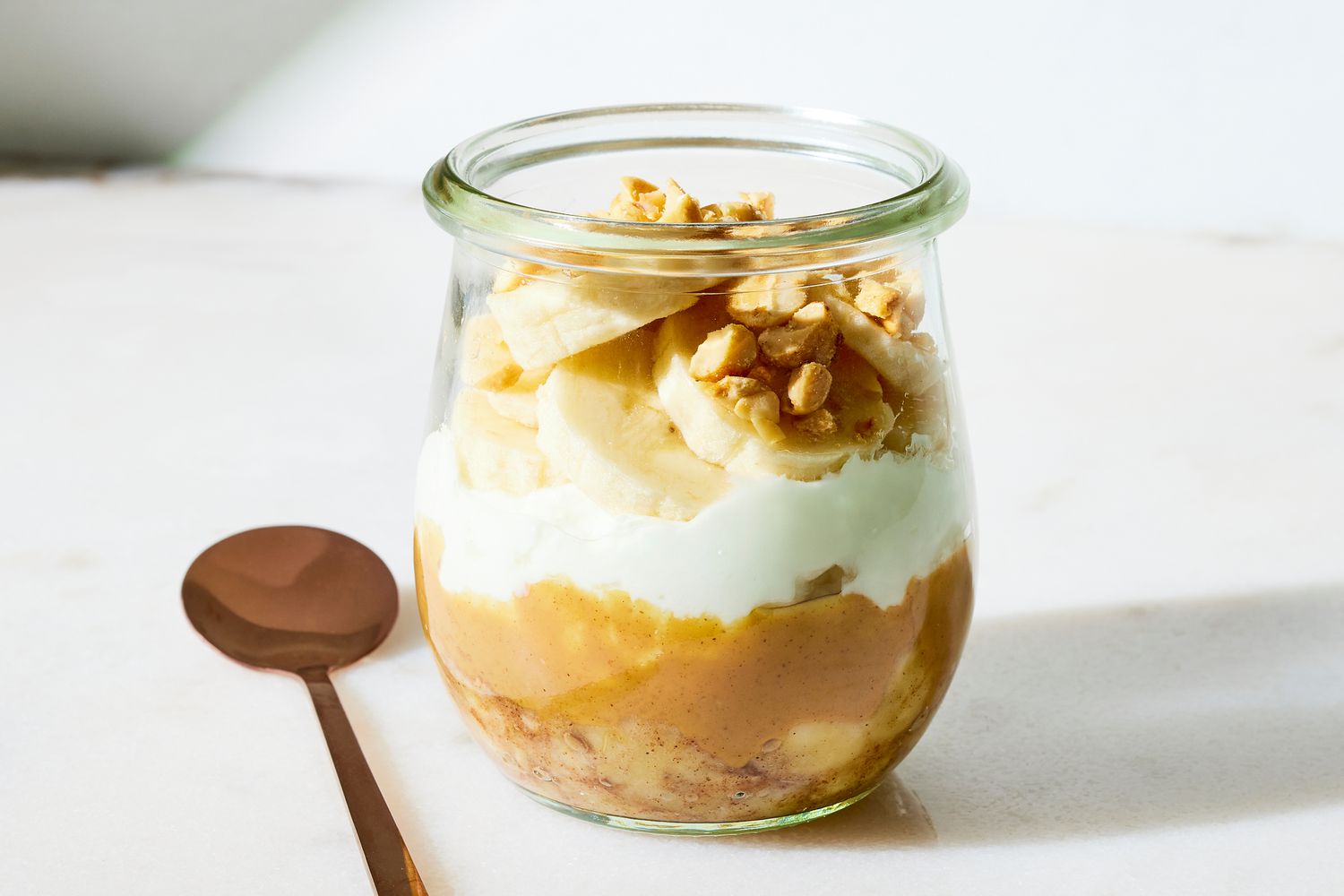 Plain yogurt, a ripe banana, and a jar of peanut butter arranged on a clean kitchen counter with a measuring spoon