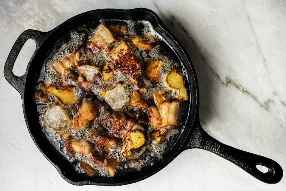 Pork belly pieces sizzling and browning in a cast iron skillet on a stovetop, with rendered fat bubbling around the meat