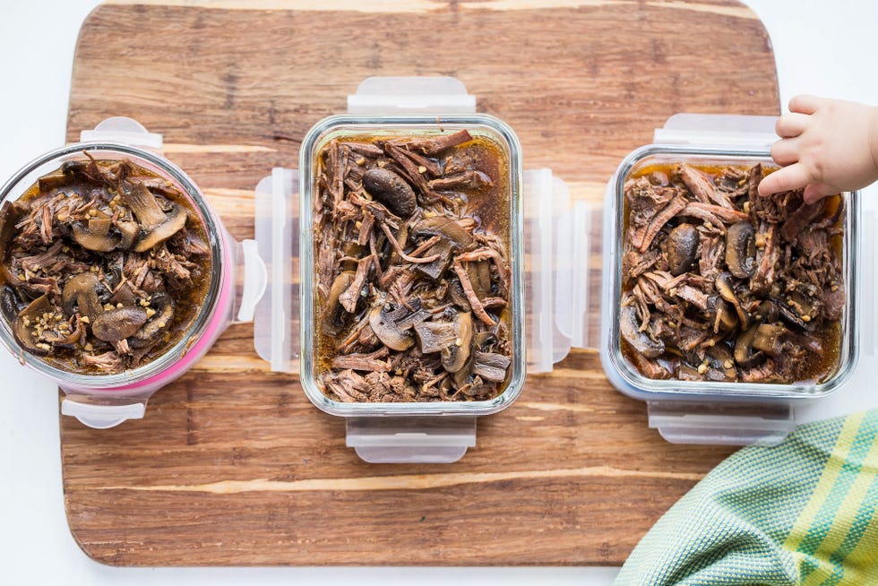 Portion containers filled with pulled beef and sauce cooling on a kitchen counter next to labels and a spoon