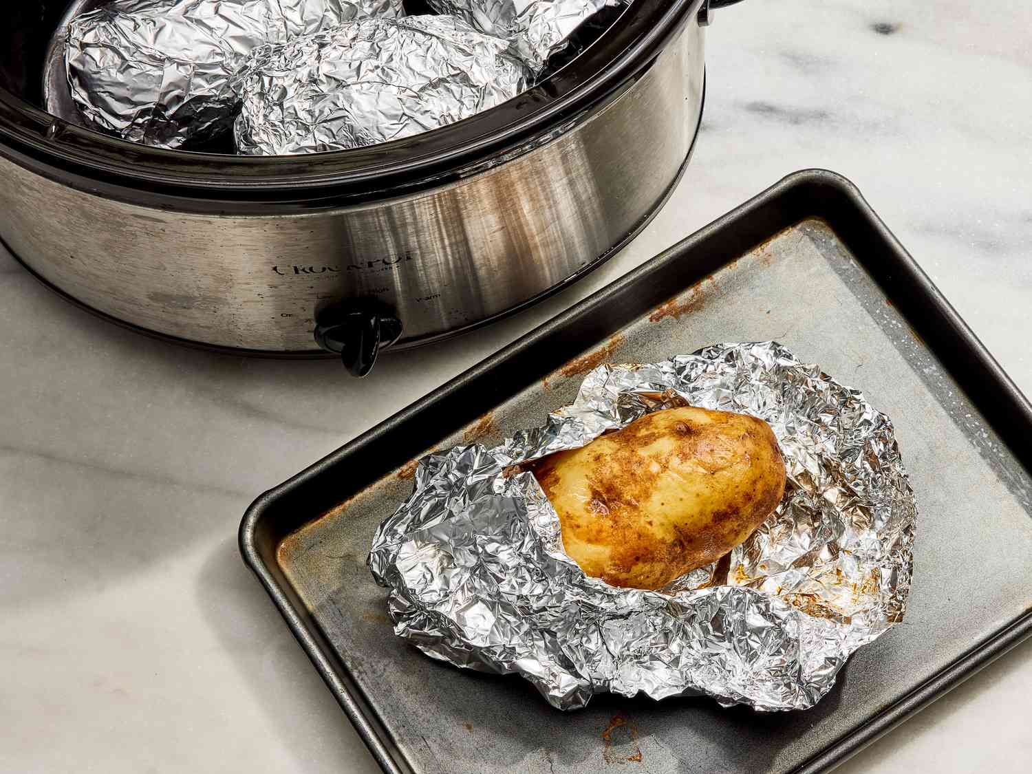 Potatoes cooking inside a slow cooker with the lid lifted, showing steam and garlic butter coating the potatoes on a neutral kitchen counter