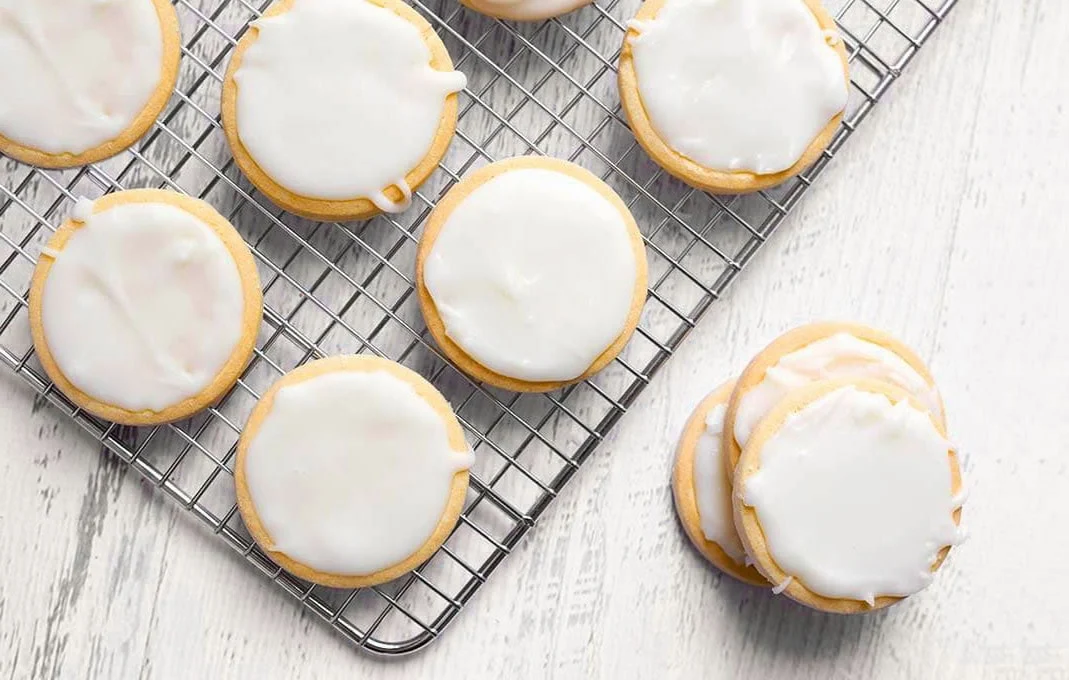 Powdered sugar, a small bowl of corn syrup, a bottle of vanilla extract, and small cups of gel food coloring arranged on a clean kitchen counter with measuring spoons