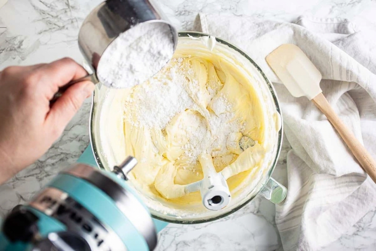 Powdered sugar being added gradually to a bowl of cream cheese and butter while a mixer runs on low, with a spatula nearby