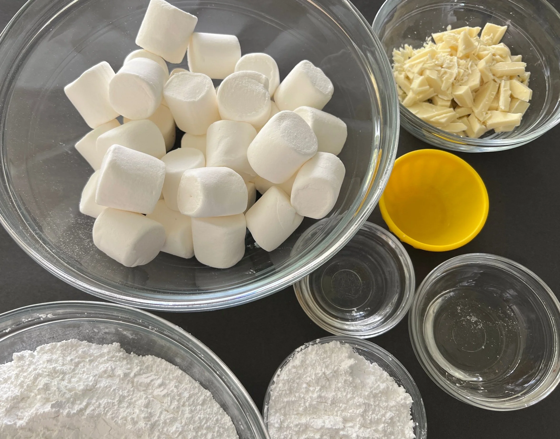 Powdered sugar, mini marshmallows, corn syrup, vanilla extract, and a bowl of water measured out on a tidy kitchen counter