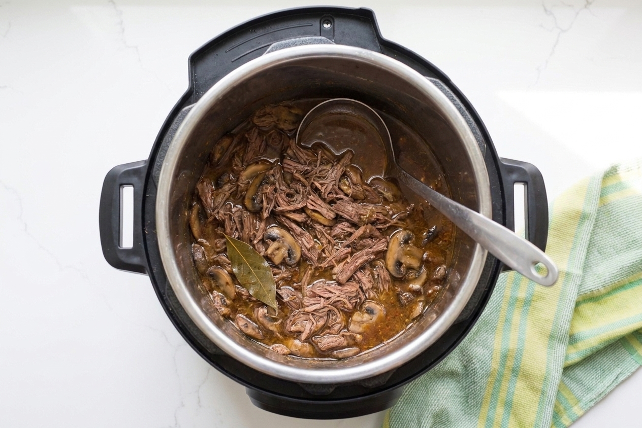 Pulled beef in an Instant Pot inner pot with sauce, bay leaf visible, and a ladle resting on the rim