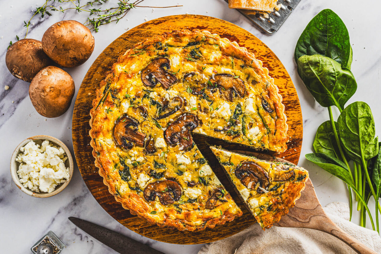 Quiche ingredients arranged on a bright kitchen counter including eggs, milk, spinach, mushrooms, bell pepper, onion, shredded cheese, and a pie crust in its pan