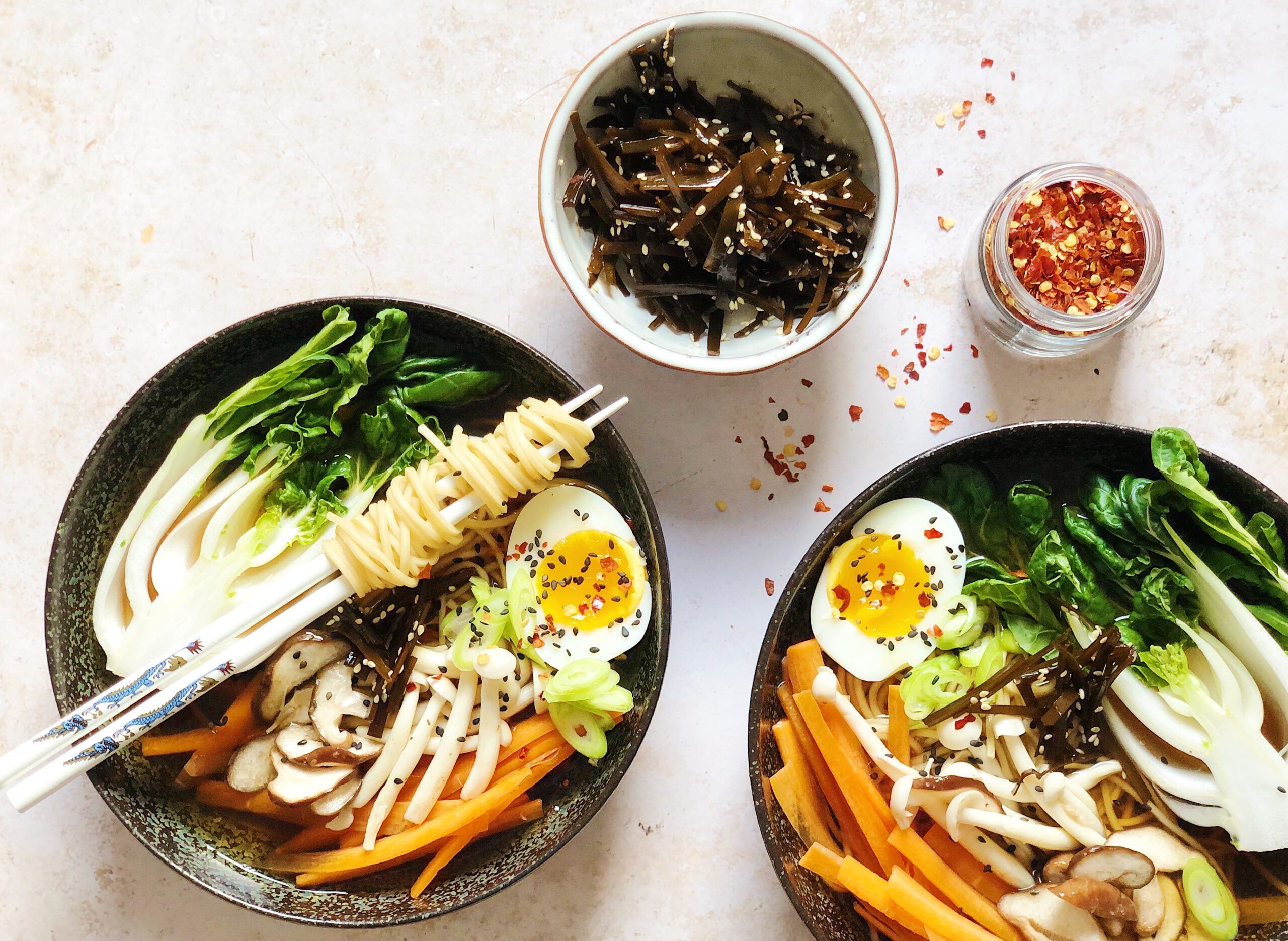 Ramen ingredients laid out on a kitchen counter including chicken bones, pork shoulder, soy sauce, mirin, ginger, garlic, scallions, dried kombu, and ramen noodles