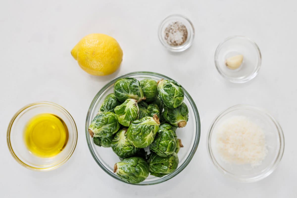 Raw Brussels sprouts, a lemon, garlic, Parmesan, olive oil, salt, pepper, and red pepper flakes arranged on a clean kitchen counter